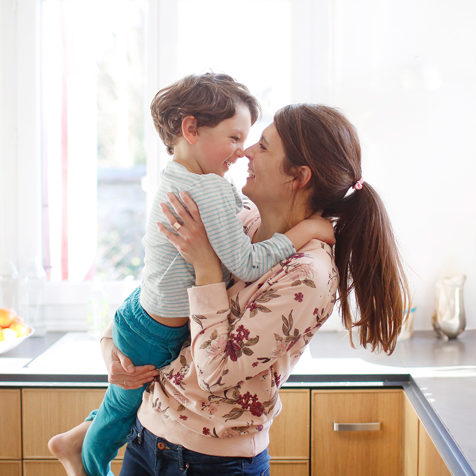 Smiling mother with brown hair in a ponytail holds her smiling son in a sunny kitchen