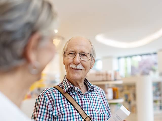 Older white man with white hair and moustache smiles and speaks with a Corewell Health pharmacy team member