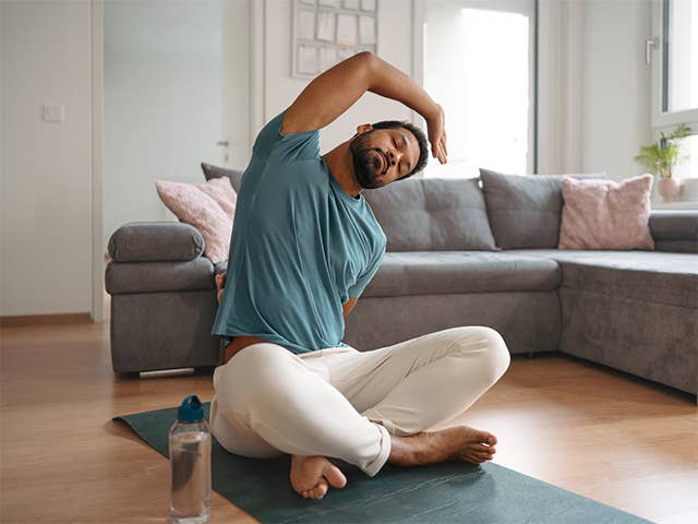 Young Black man sitting on a yoga mat in his living room and stretching his body to his left