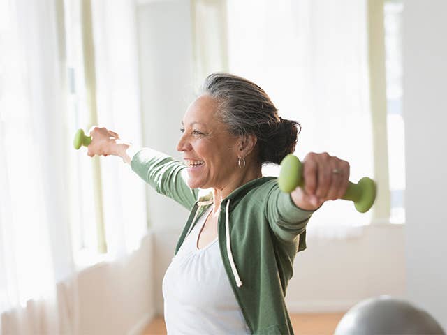 Smiling woman wearing a white shirt and green jacket holds dumbbells in her hand as she stretches her arms out to her sides