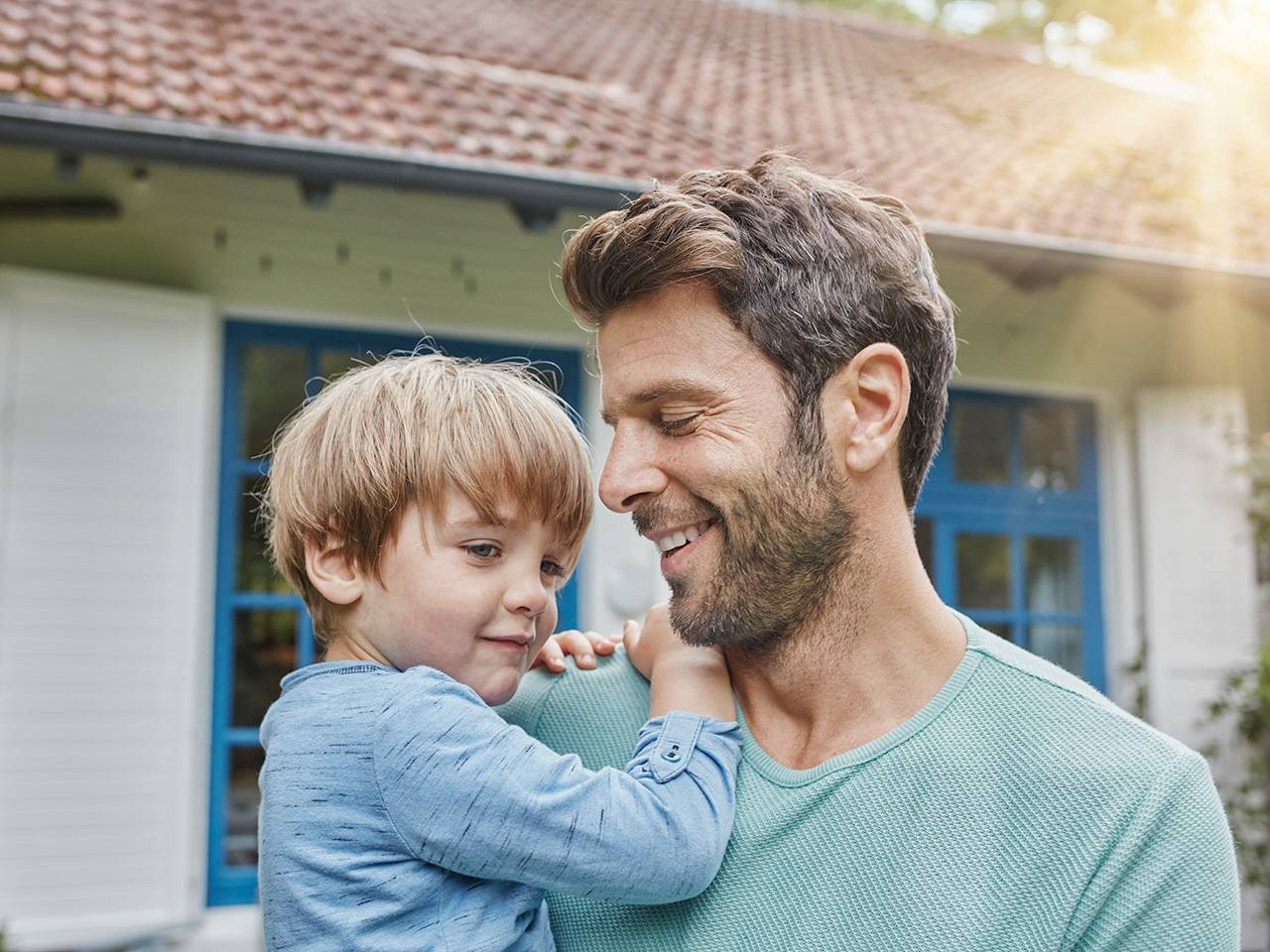Smiling father with brown hair and a beard stands outdoors in front of a house holding his young son