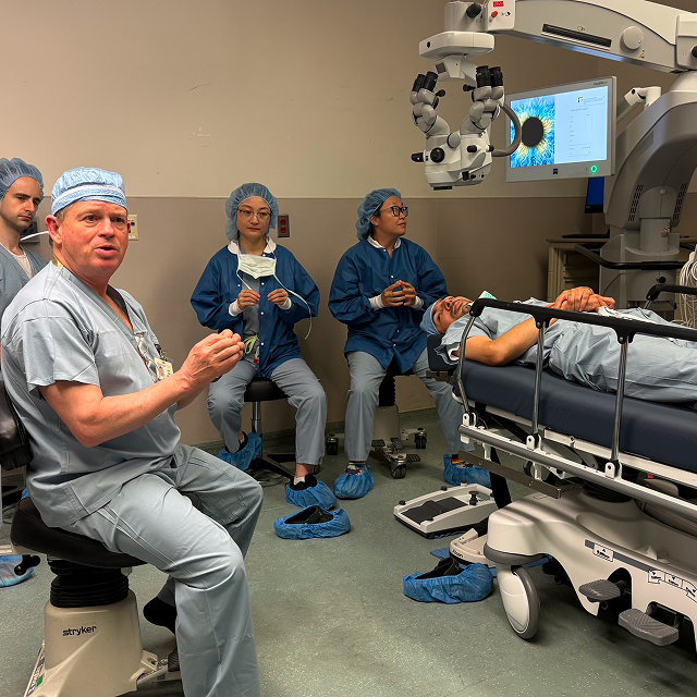 Medical instructor demonstrates an eye procedure while trainees observe beside a patient in an operating room.