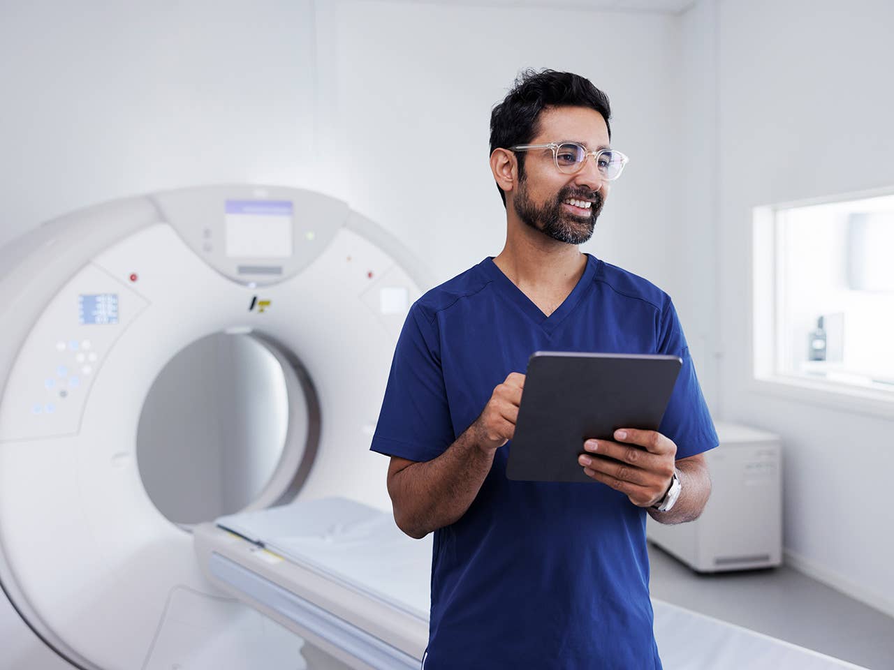 Man with glasses and a beard and wearing blue scrubs stands in front of an MRI machine