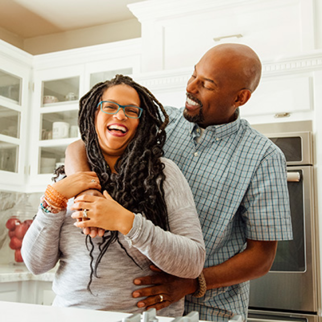 Smiling Black man stands behind smiling Black woman as they hug