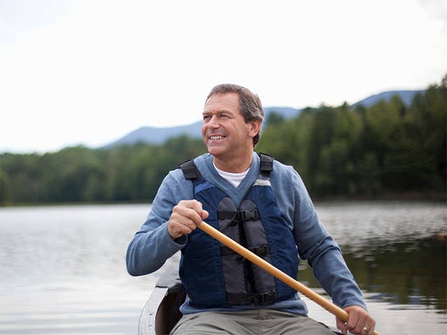 Older white man wearing a navy blue life jacket and holding an oar sits in a boat on a lake