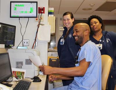 Three medical professionals smiling and interacting at a hospital workstation, with computer screens and medical equipment visible in the background.