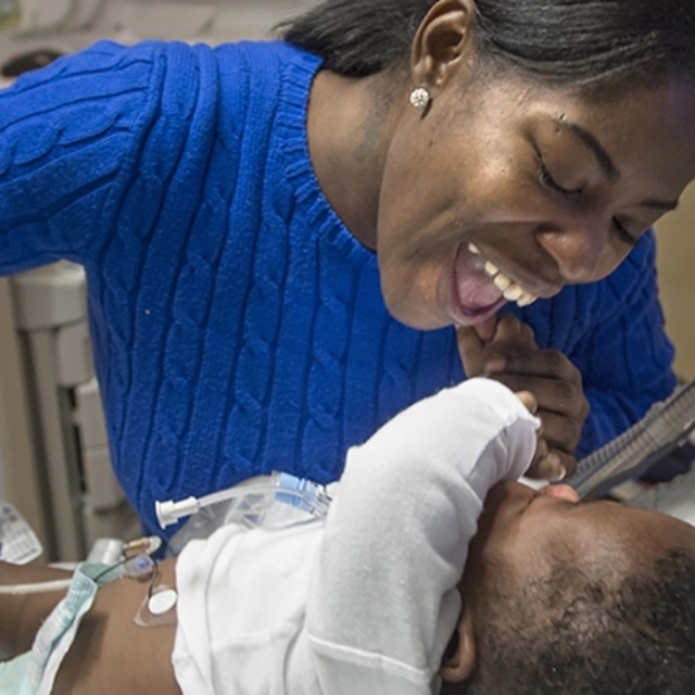 Young Black woman in a blue shirt smiles at her baby in the neonatal intensive care unit