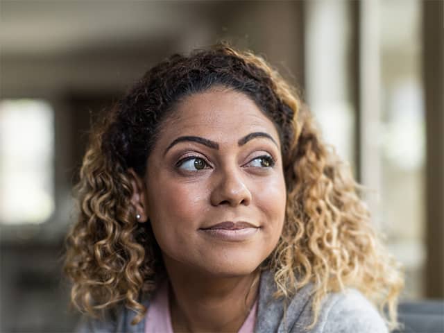 Black woman wearing a pink blouse and grey sweater sits in a sunlit kitchen