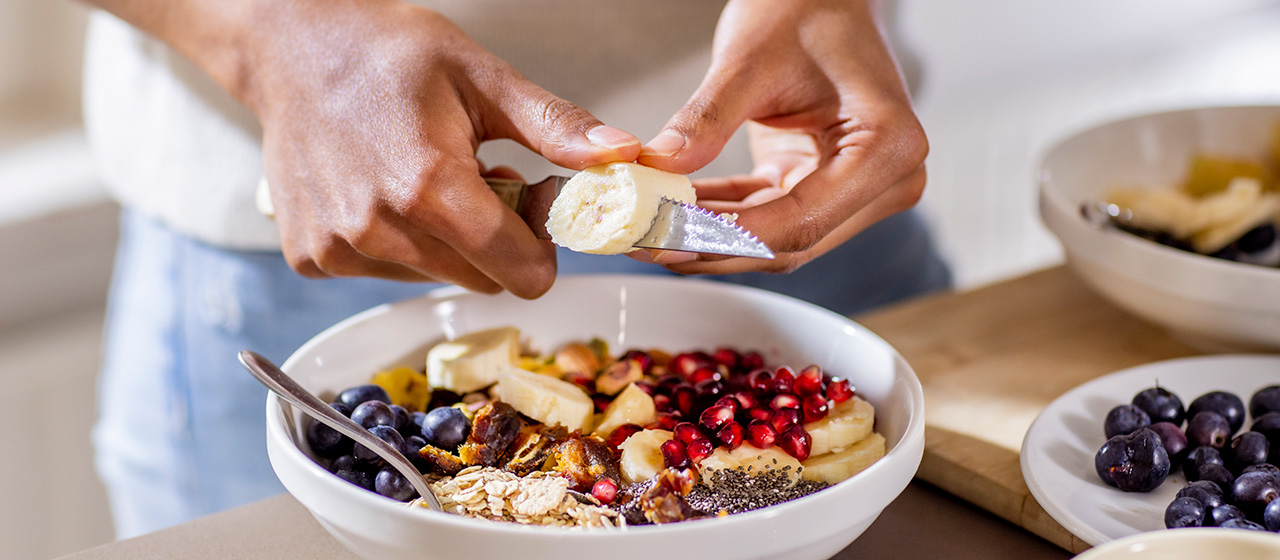Hands slicing banana over a breakfast bowl with berries, oats, chia seeds and pomegranate seeds on a kitchen counter.