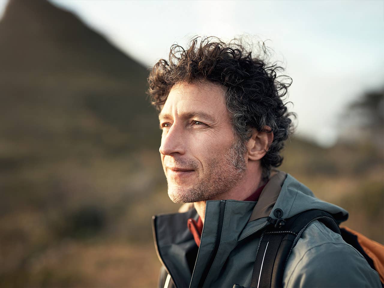 Middle aged white man with curly brown and grey hair stands in the evening sun with a mountain in the background