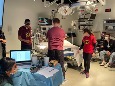 A group of individuals in a hospital room collaborates using a laptop, engaged in pediatric emergency simulation training.