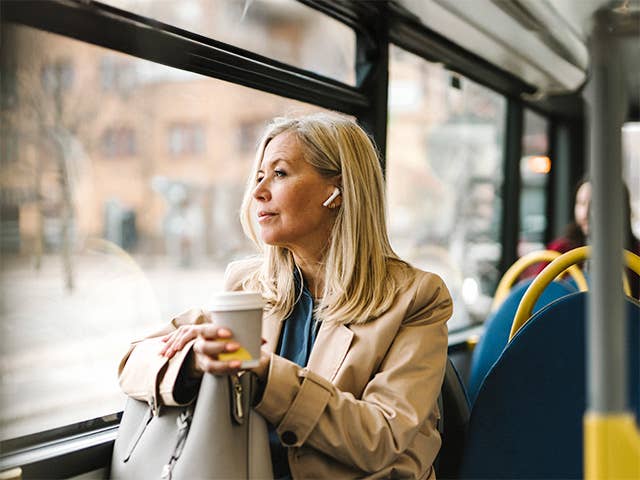 Middle aged white woman sits on a bus while wearing earbuds