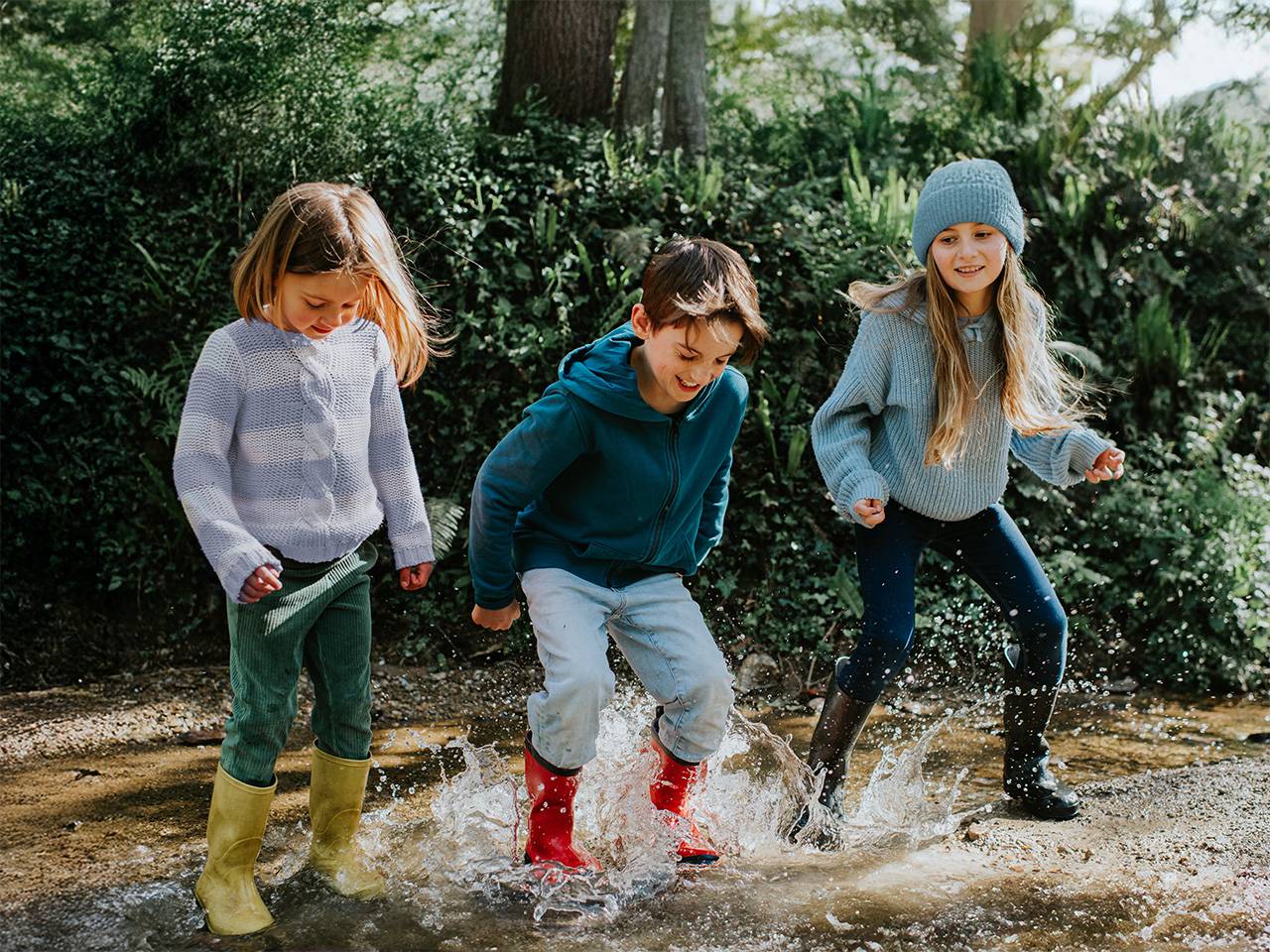 A boy and two girls jump and splash in a puddle in the sunshine outdoors
