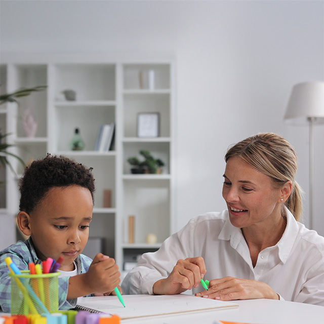Smiling woman wearing a white blouse sits next to a young Black boy at a table as they color with markers