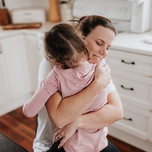 Smiling mother with brown hair and wearing a white shirt hugs her young daughter in the kitchen