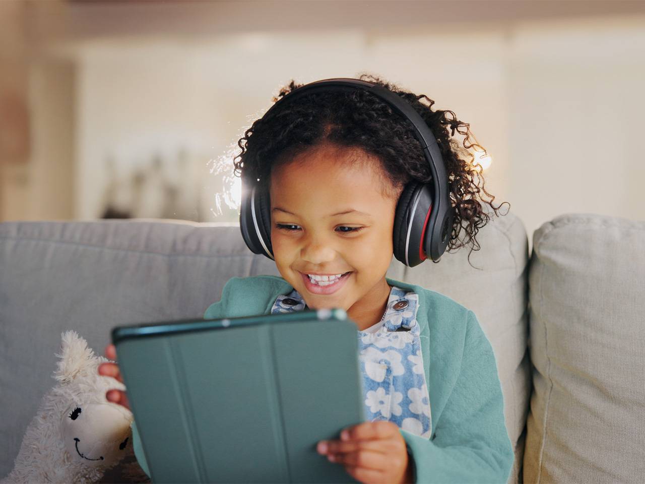 A young Black girl with curly brown hair smiles and looks at a tablet device while wearing headphones