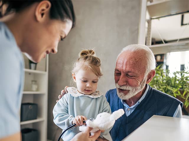 Child using a stethoscope on a stuffed animal while seated with an older adult as a healthcare worker assists.