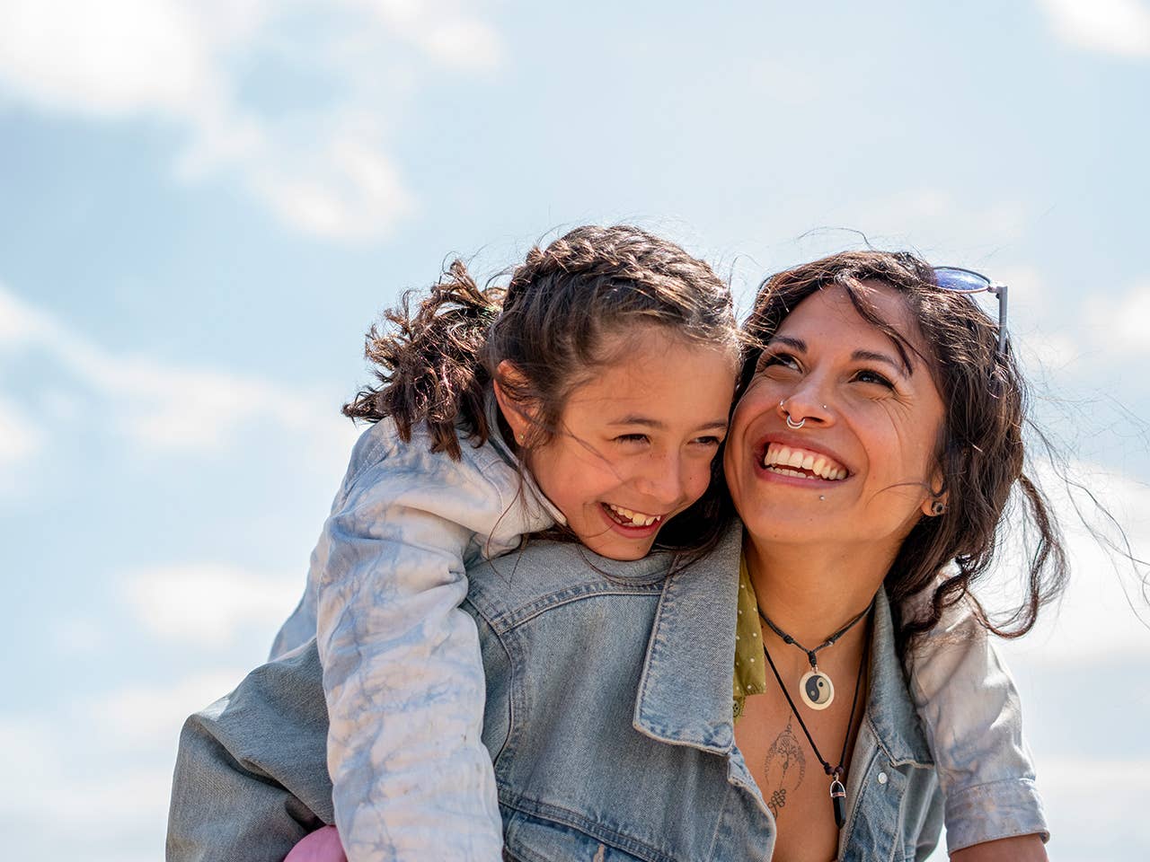 Smiling woman wearing a denim jacket carries her young daughter on her back outdoors in the sunshine