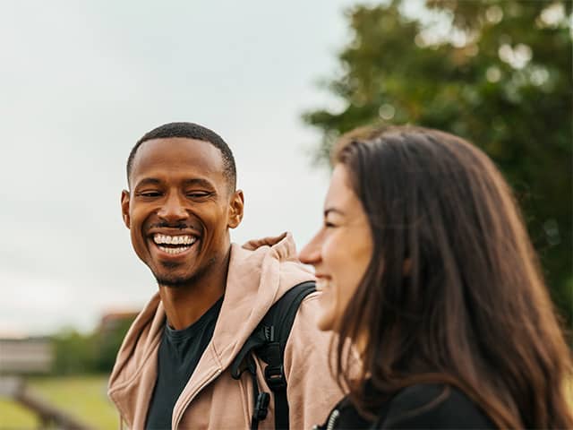 Young Black man wearing a light brown jacket smiles and laughs while talking with a woman outdoors