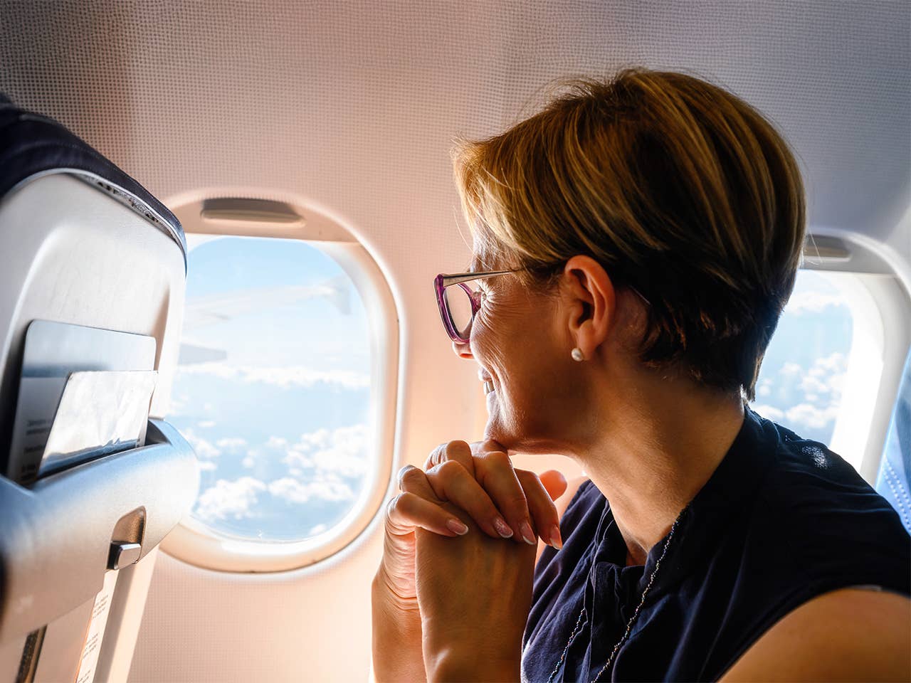 A woman peers out the window of an airplane, watching the clouds and scenery as she travels through the sky.