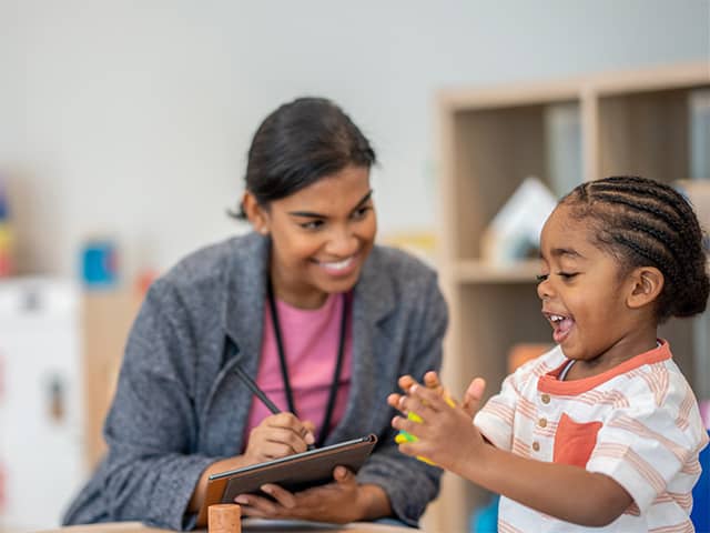 Smiling Black girl claps her hands while seated next to a smiling Corewell Health child life specialist