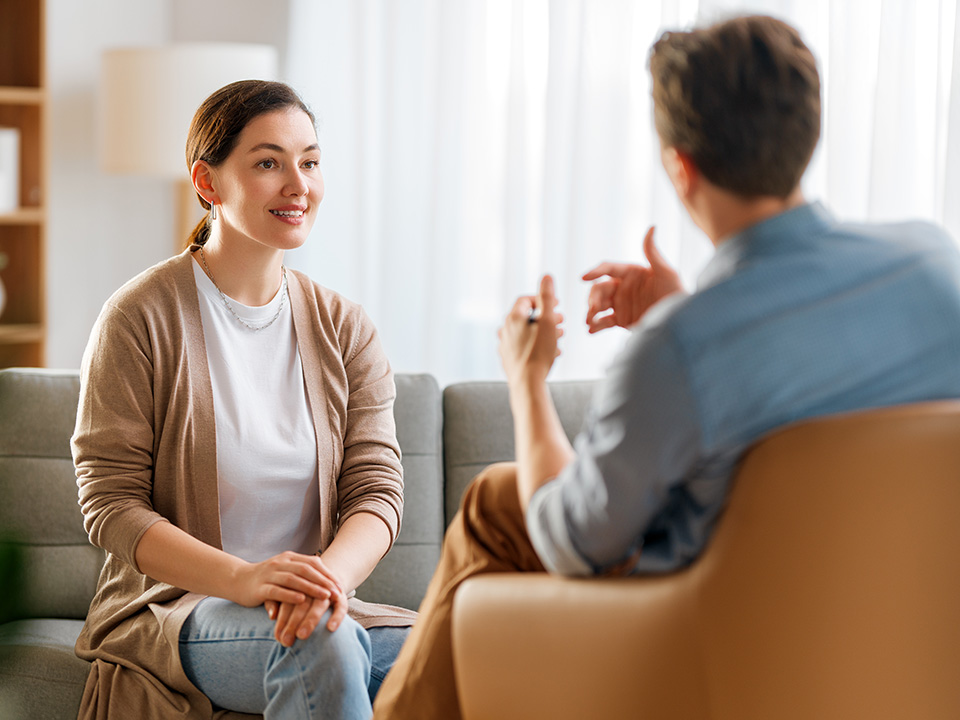 A man and woman on a couch, talking and sharing a light moment, with relaxed body language and friendly expressions.