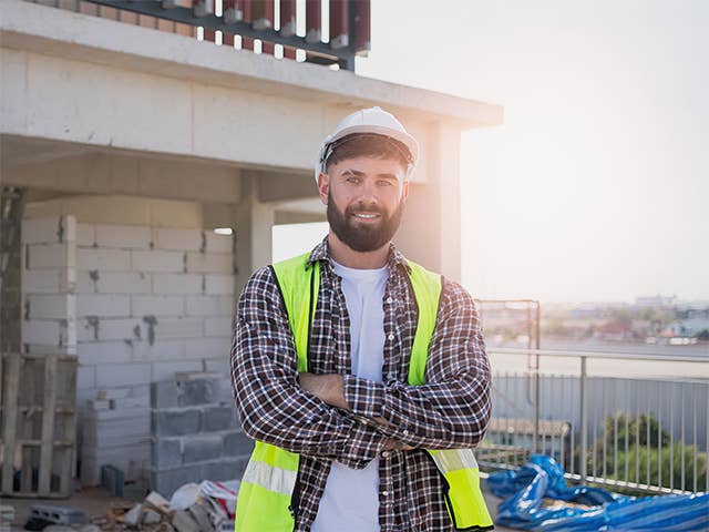 Smiling white man wearing a neon yellow safety vest and construction helmet stands on a work site in the morning sun