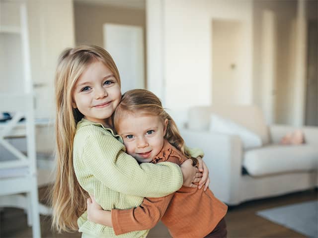 Two young girls with long blonde hair smile and hug each other in a living room