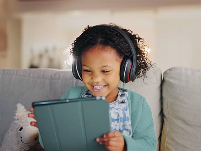 A young Black girl with curly brown hair smiles and looks at a tablet device while wearing headphones