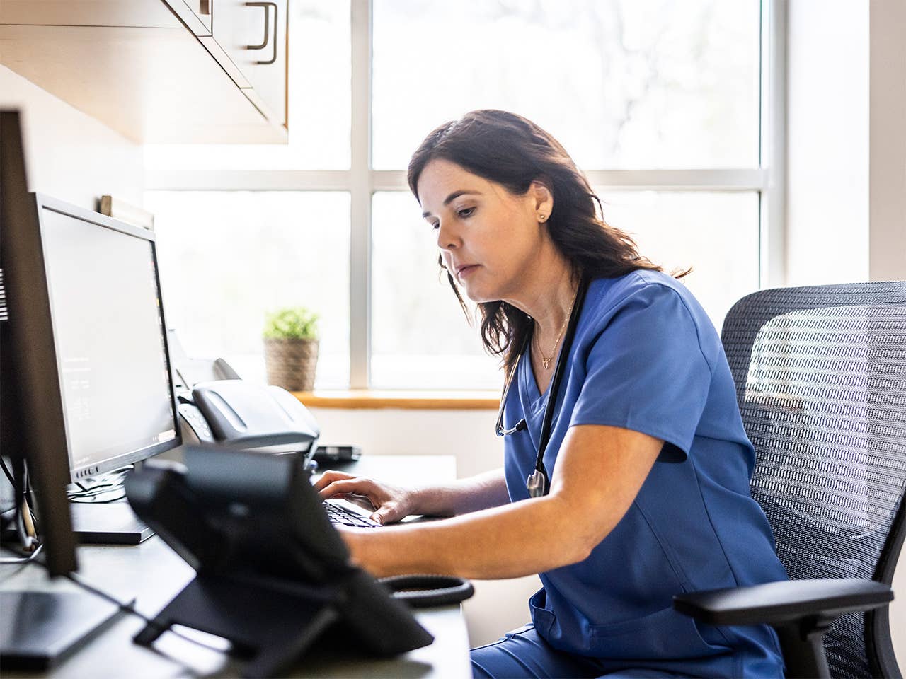A woman in scrubs seated at a desk, focused on her work while using a computer.