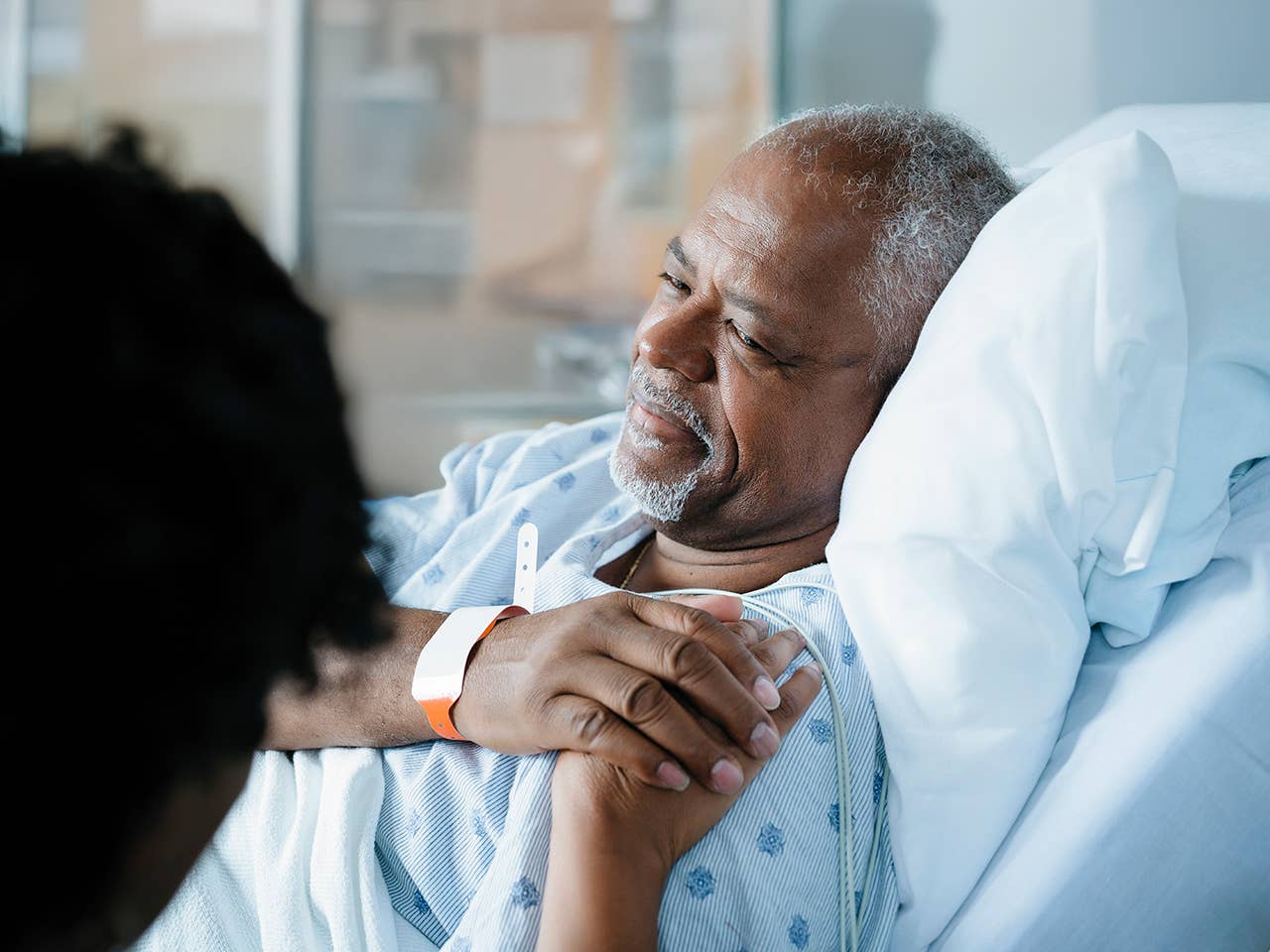 Older Black man laying in a hospital bed smiles and holds hands with his wife