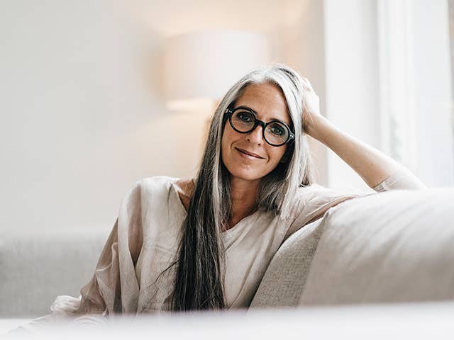 Smiling older white woman wearing glasses rests her elbow on a couch