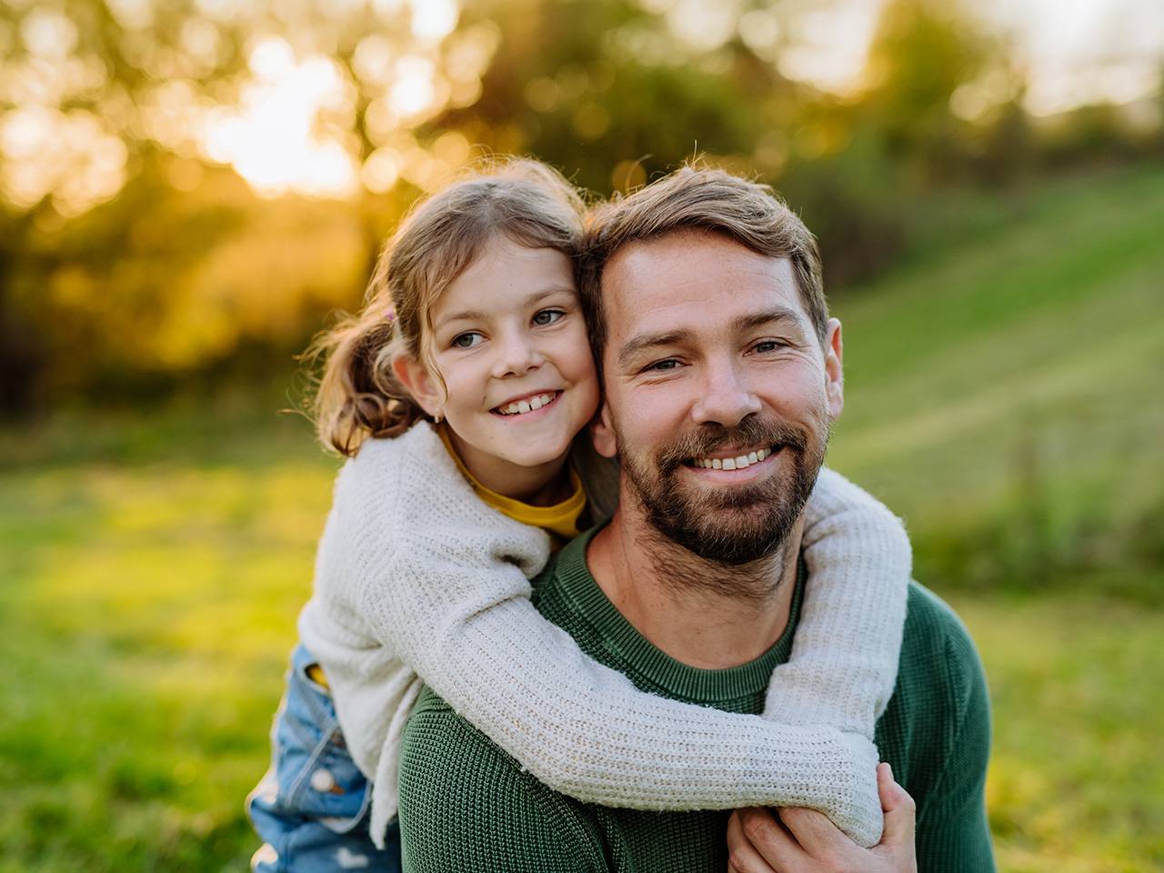 Smiling man with brown hair and a beard carries his smiling daughter on his back while they are outdoors in the sunset