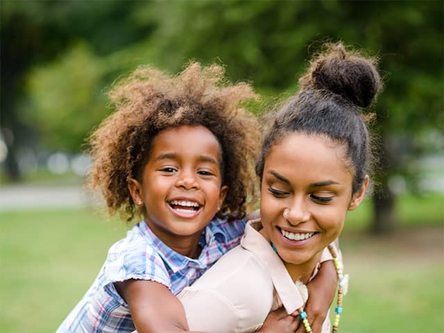 Smiling Black woman looks back at her smiling daughter while carrying her on her back outdoors
