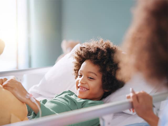 A young Black boy with curly brown hair lies in a bed inside a sunlit room