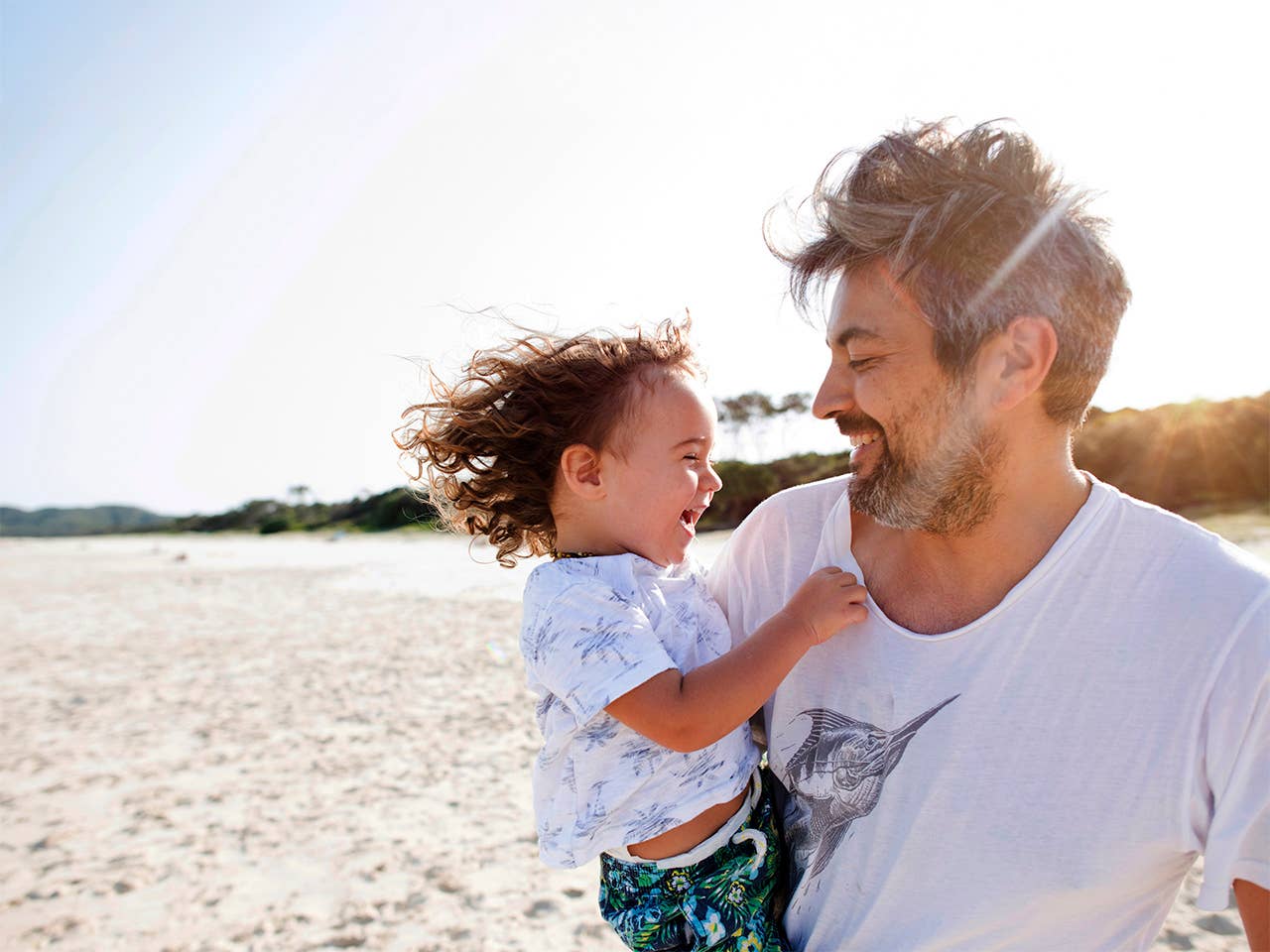 Middle-aged white man holds his toddler son while standing in the sunshine on the beach
