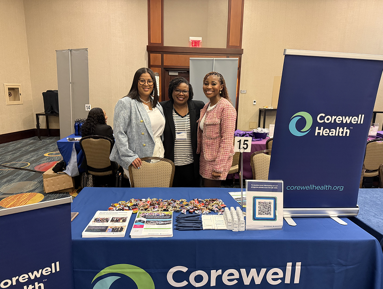 Three individuals standing behind a Corewell Health display table at an indoor event, with brochures, candy, and branded items on the table, and a large Corewell Health banner in the background.