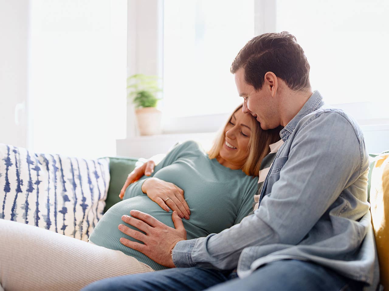 Young man hugs his pregnant wife and places a hand on her belly as they sit on a couch