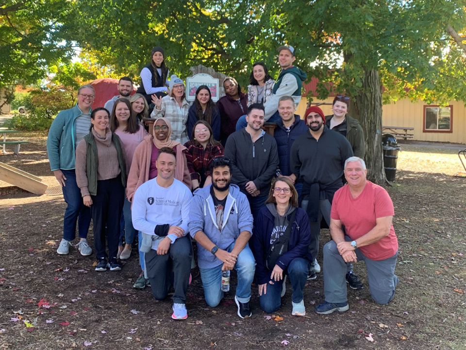 A large group of people posing together outdoors at a fall gathering, standing and kneeling beneath colorful trees with a barn and picnic area in the background.