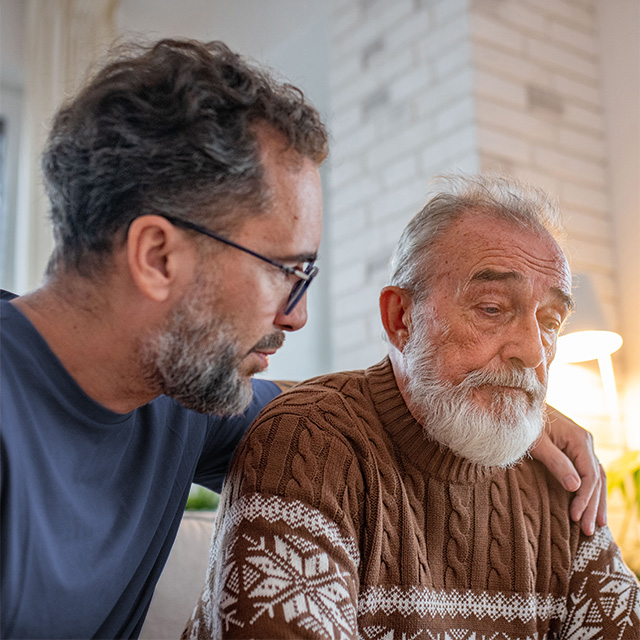 Adult sitting beside an older adult with a hand on the shoulder in a home setting, offering support and companionship.