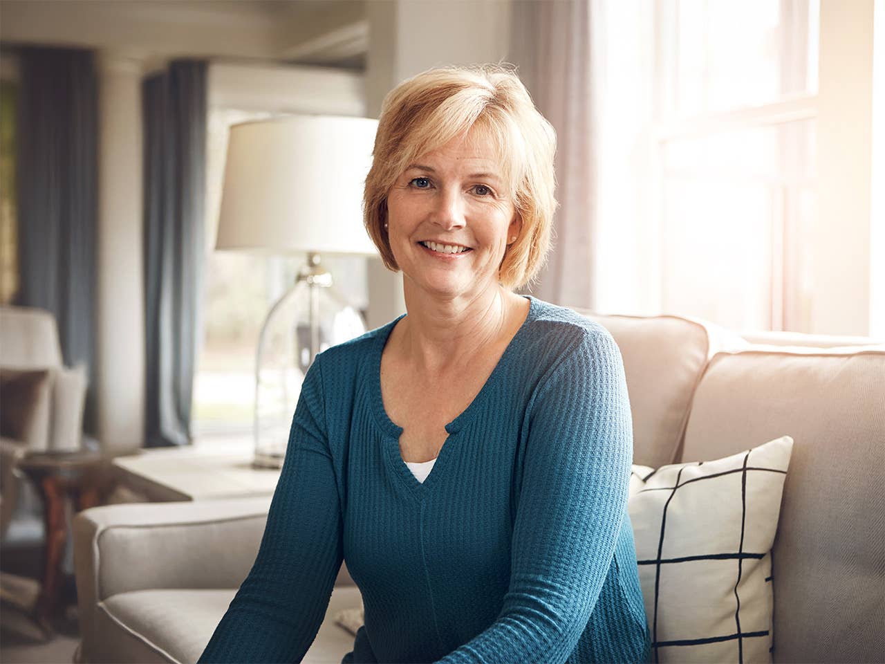 White woman in a teal sweater smiles while she sits in a sun filled room