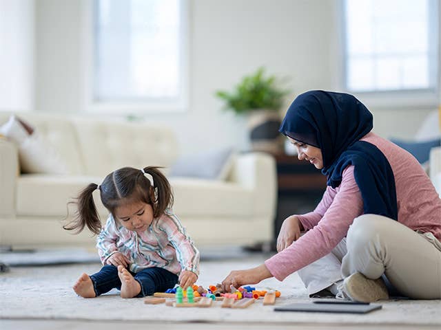 Smiling woman sits on the floor next to her young daughter, playing with toys