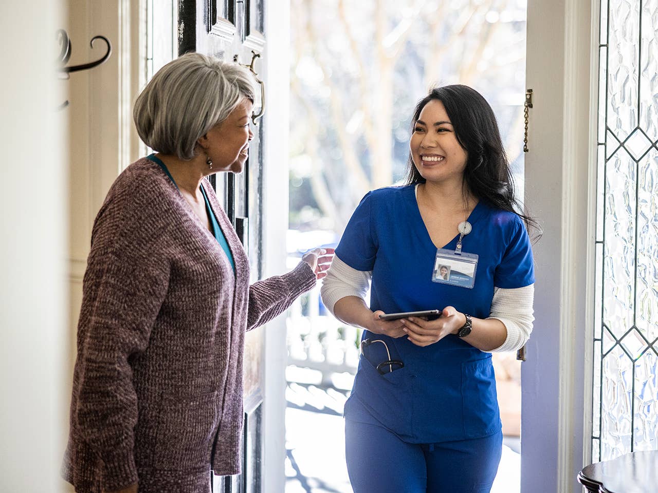Smiling Corewell Health home based primary care provider wearing blue scrubs speaks with an elderly woman inside her front door