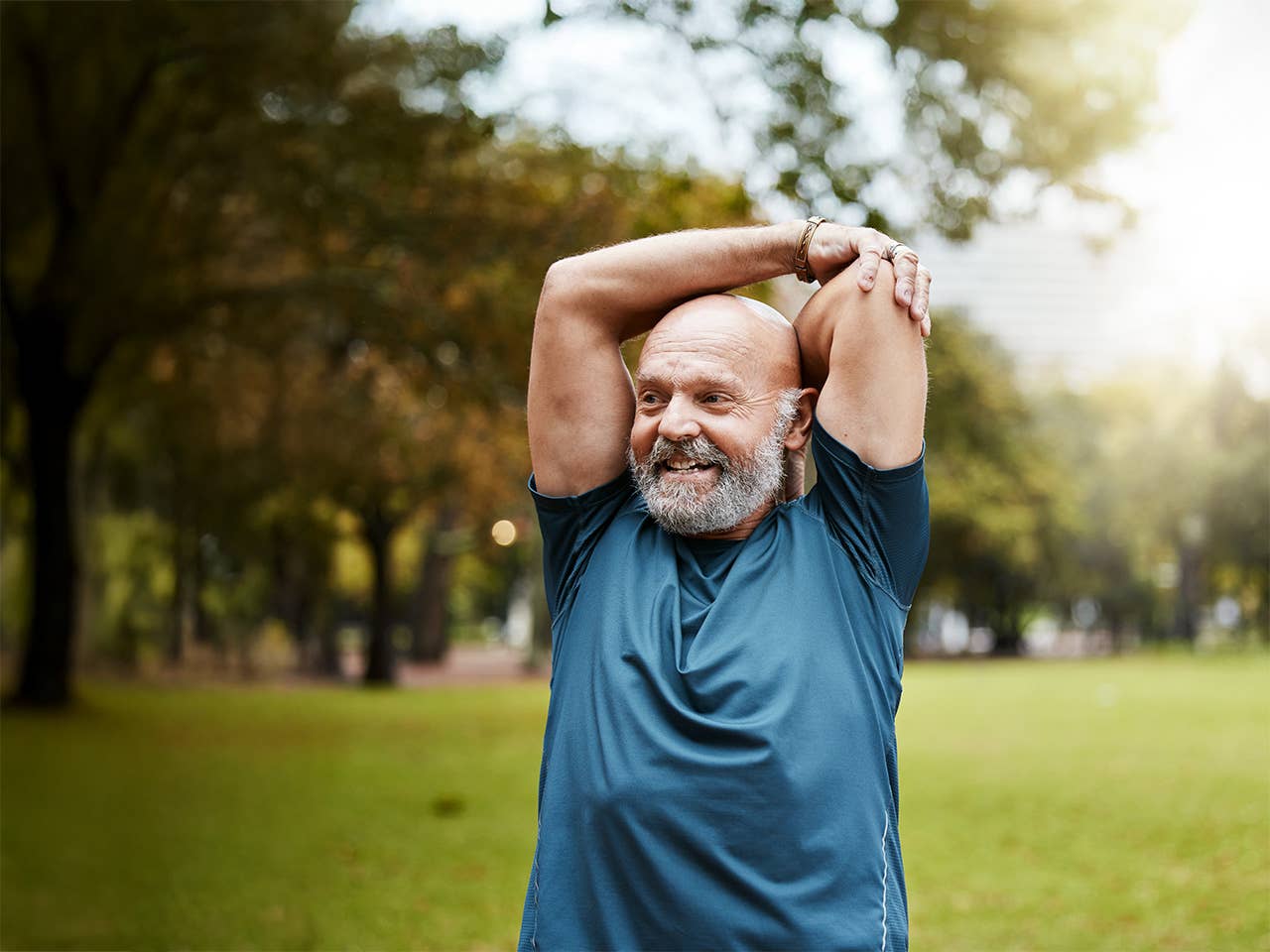 White man with a bald head and grey beard stretches his arms over his head while standing outdoors