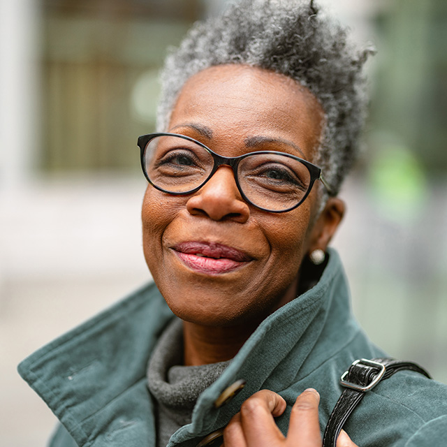 An older woman wearing glasses and a green jacket smiles warmly.
