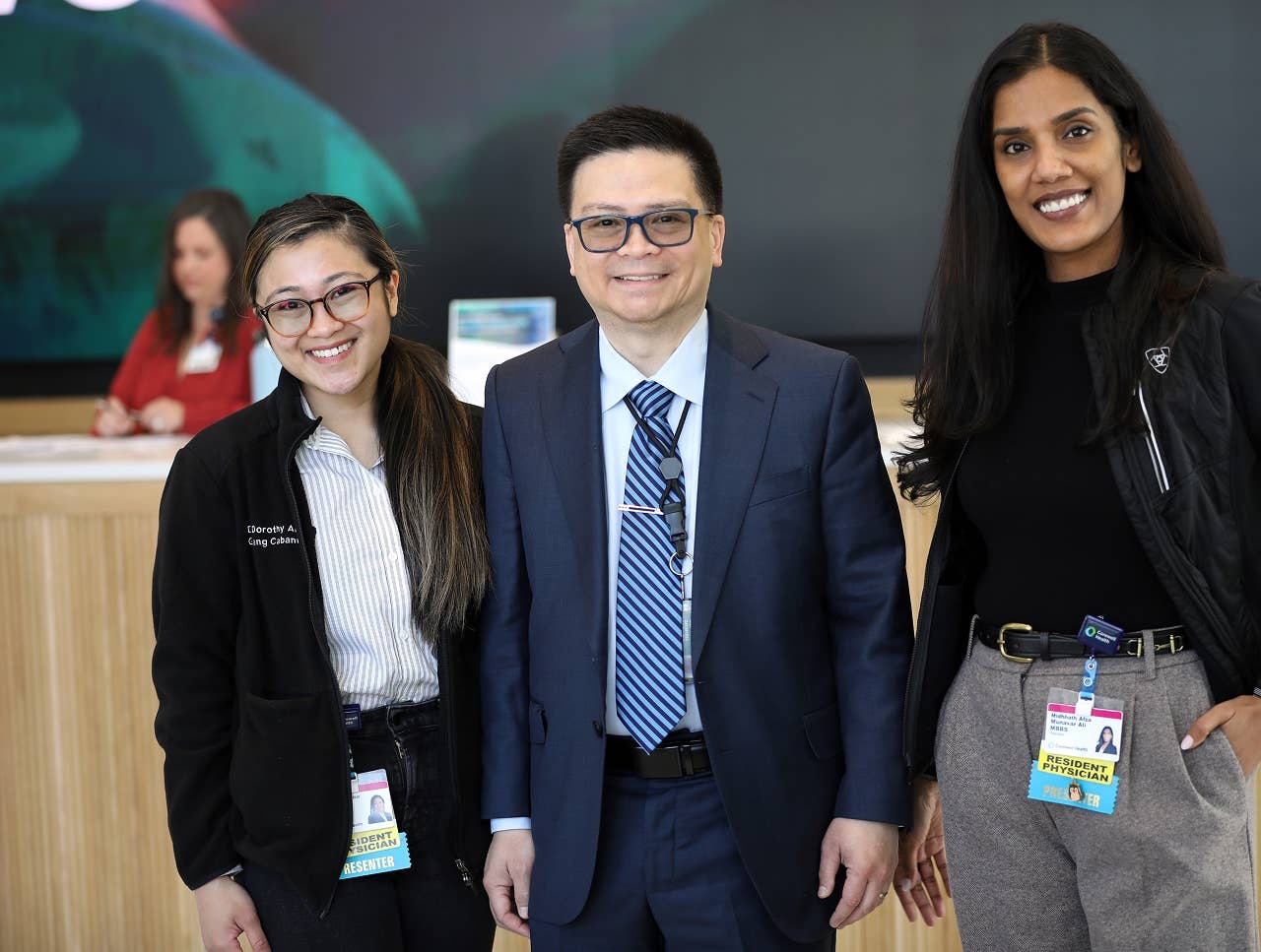Three Corewell Health team members standing together in a modern lobby area, wearing professional attire and ID badges, with a reception desk and digital display in the background.
