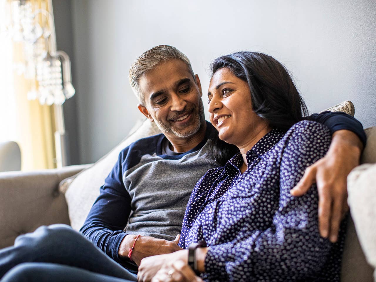 Smiling middle aged Indian man and woman sit beside one another on a couch hugging