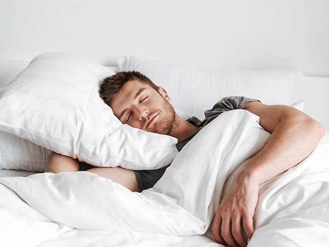 Man with brown hair and a beard sleeps on his side in a bed with white sheets and blankets