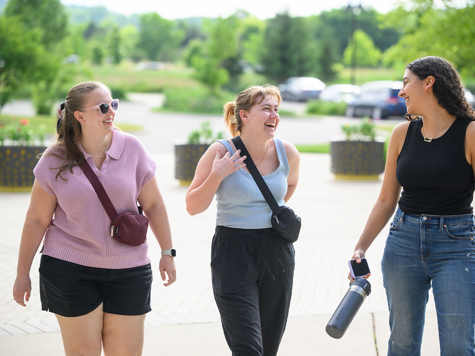 Three people walking together outdoors on a sunny day, talking and smiling as they walk along a paved path with greenery and planters in the background.
