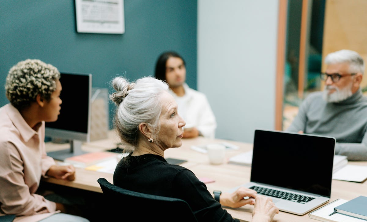 Mulher mais velha com cabelos brancos atenta com sua equipe durante uma reunião sobre habilidades profissionais e resolução de conflitos.